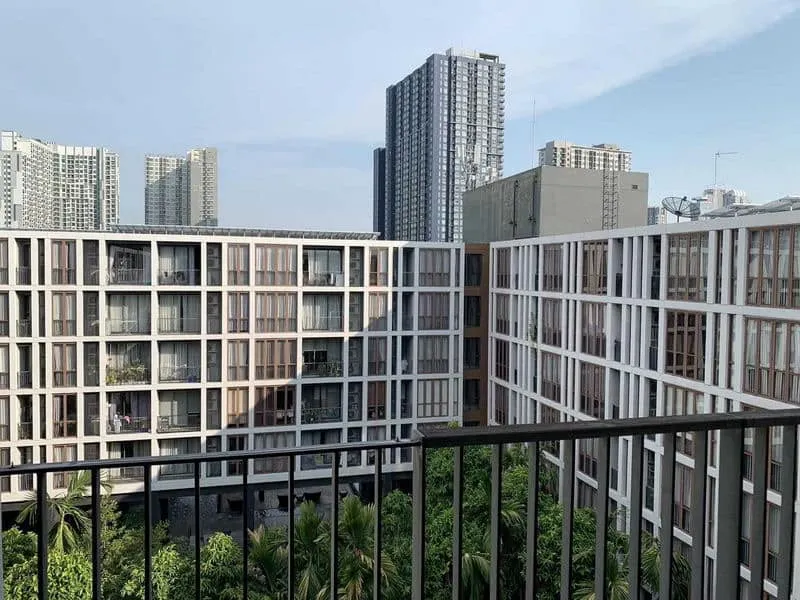 Modern apartment buildings with large windows surround a courtyard with green trees, set against a backdrop of high-rise skyscrapers under a clear sky.