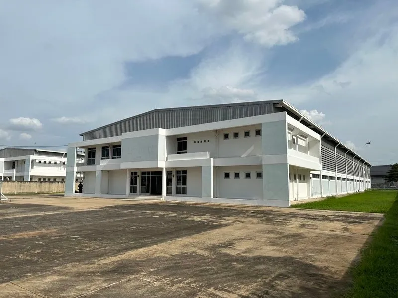 A large two-story white industrial building with a metal roof, surrounded by a concrete yard and grassy area under a partly cloudy sky.