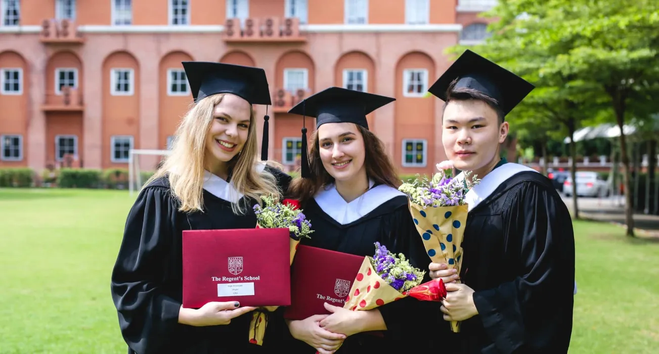 บัณฑิตในชุดครุยและหมวกรับปริญญา ยิ้มแย้มแจ่มใสพร้อมใบปริญญาและดอกไม้ ยืนอยู่หน้าอาคา