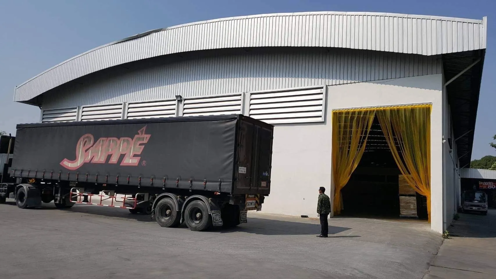 โกดังขนาดใหญ่ มีรถบรรทุกจอดอยู่ด้านนอก มีม่านโปร่งแสงสีเหลืองกั้นอยู่ที่ทางเข้า และมี