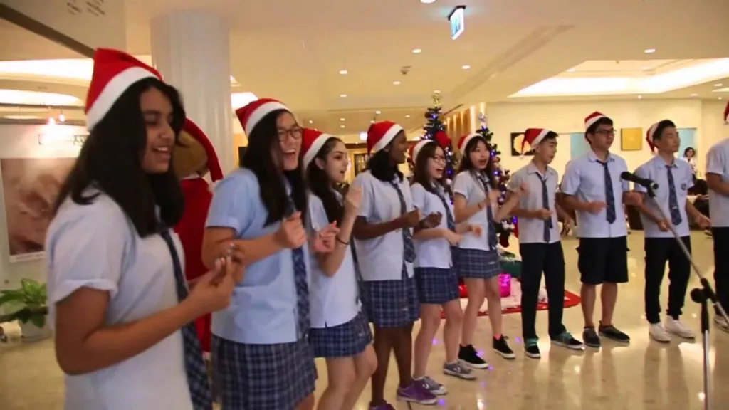 A group of students wearing Santa hats, singing and performing indoors, with Christmas decorations and a tree in the background.