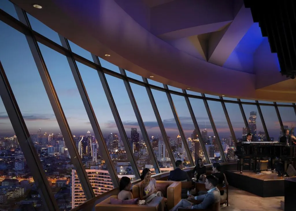 Sky lounge with people seated, enjoying a panoramic city skyline view at dusk through large windows, with a pianist and singer performing.