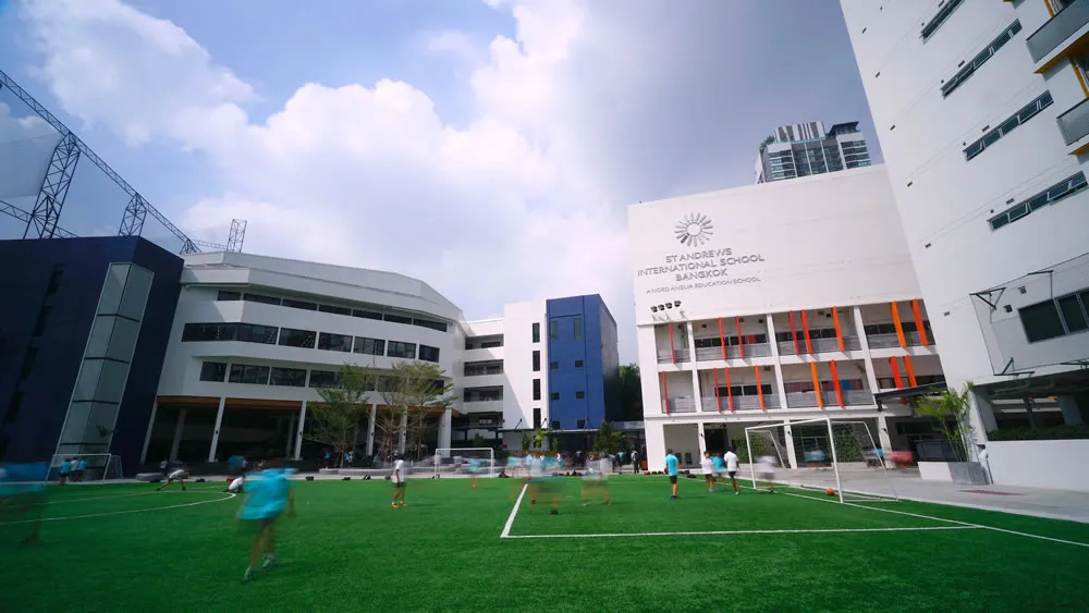 ภาพโรงเรียนสมัยใหม่ที่มีนักเรียนกำลังเล่นอยู่บนสนามหญ้าสีเขียว ล้อมรอบด้วยอาคารสีขาว