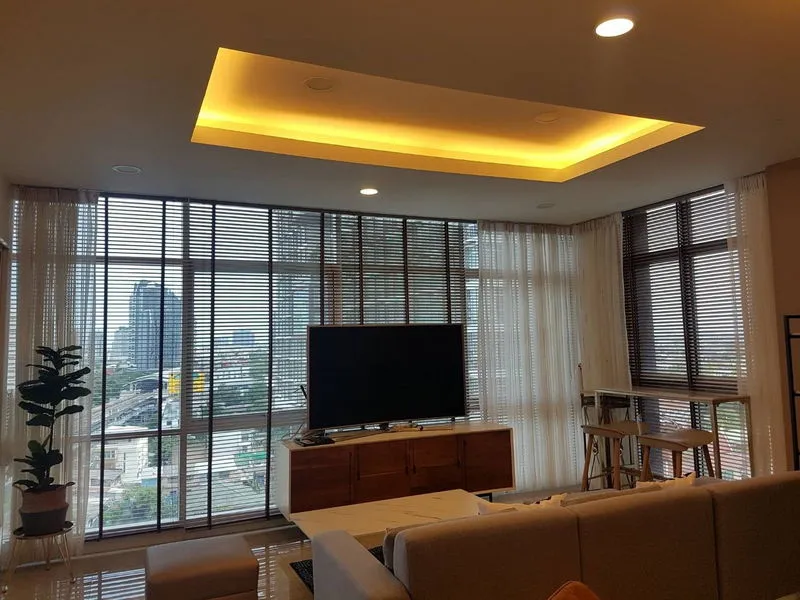 Modern living room with large windows, sectional sofa, flat-screen TV on a wooden stand, potted plant, and city view.