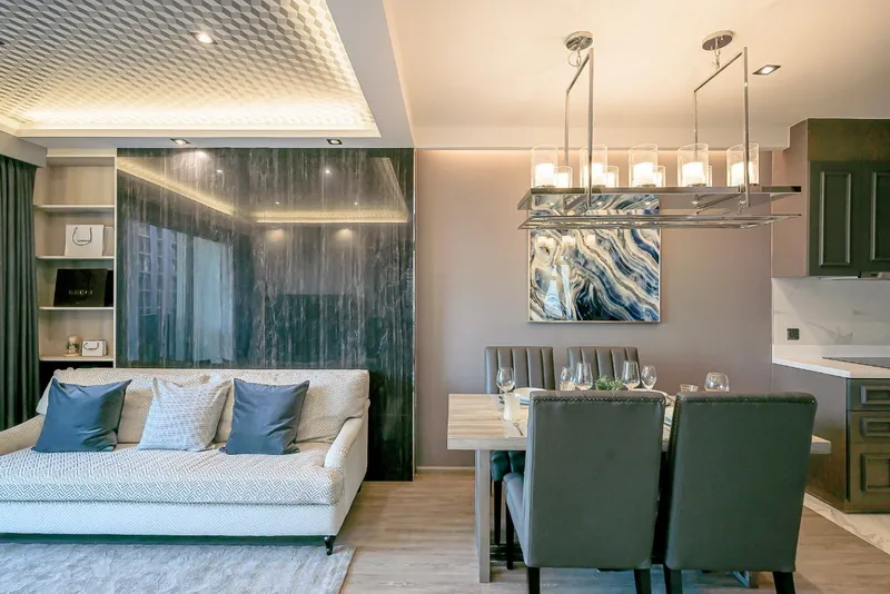 Modern living room and dining area with a plush sofa, decorative panels, abstract wall art, and a candle-style chandelier over the dining table.