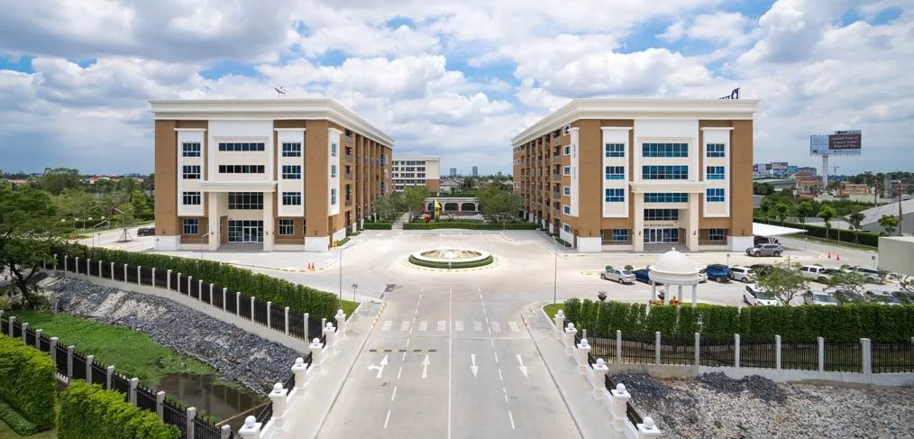 Aerial view of two modern buildings with a circular driveway and parking area, surrounded by green landscaping and a blue sky with clouds.