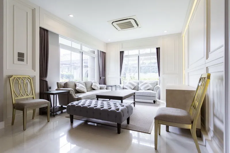 Modern living room with large windows, beige sofas, a tufted ottoman, and elegant molding. Bright and spacious with abundant natural light.