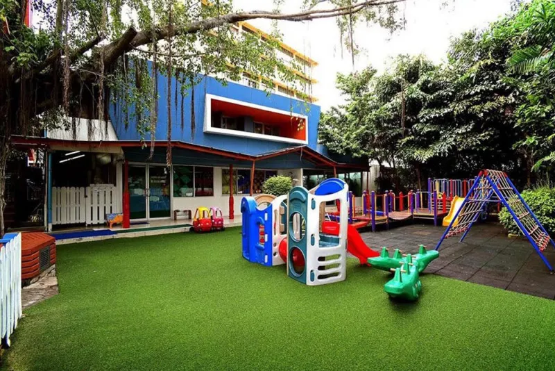 Colorful playground with slides and climbing structures on artificial grass outside a modern building, surrounded by trees.