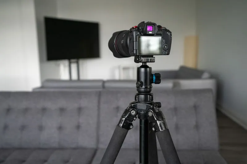 Camera on a tripod in a living room with a TV, sofa, and table in the background.