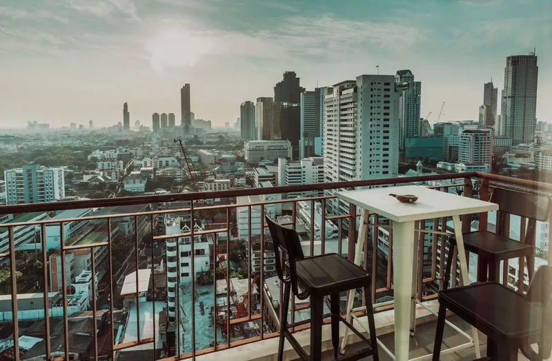 Cityscape with tall buildings viewed from a balcony, featuring a table and chairs in the foreground.