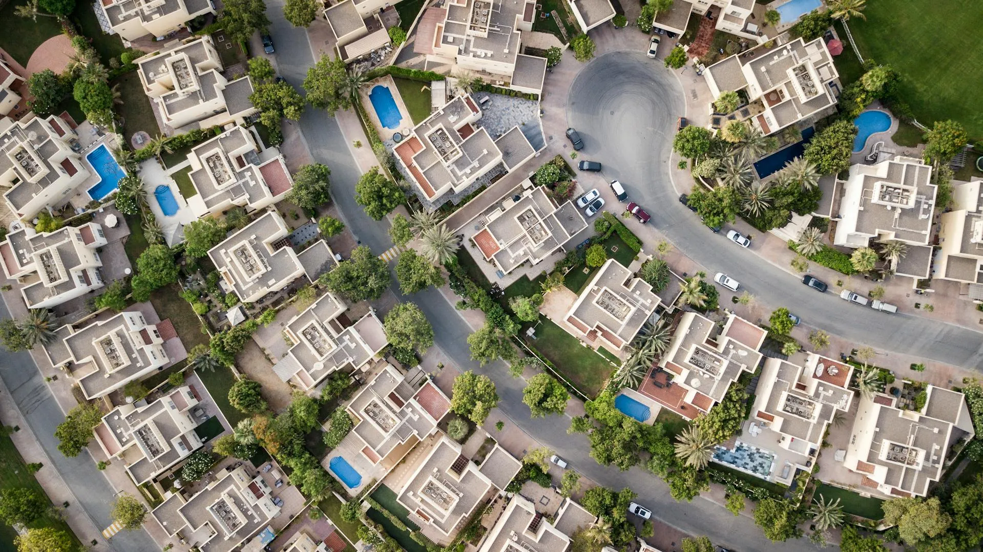 Aerial view of a suburban neighborhood with modern houses, trees, and swimming pools arranged along curved streets.