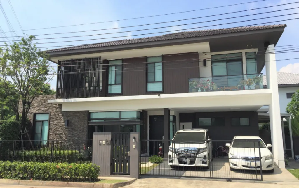 Modern two-story house with a gray roof, balcony, and two cars parked in the driveway, surrounded by a black fence and greenery.