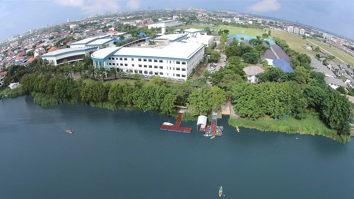 Aerial view of a campus with modern buildings, surrounded by greenery, near a river with small boats and kayaks.