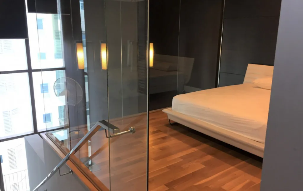 Modern bedroom with wooden floor, enclosed by glass walls, featuring a white bed and warm lighting, adjacent to a window and staircase.