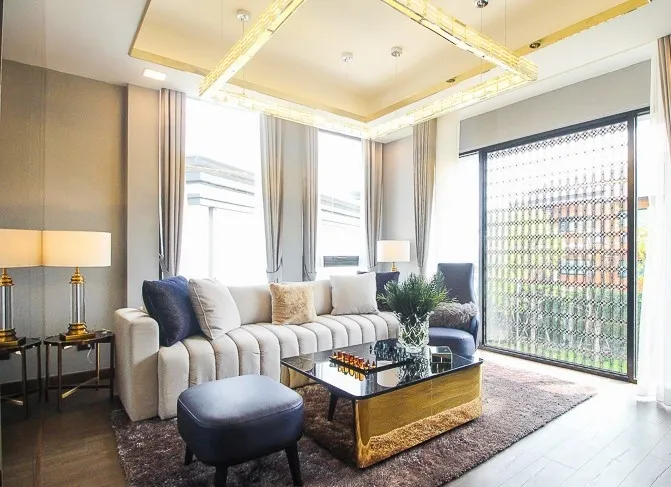 Modern living room with plush white sectional sofa, blue armchairs, gold-accented coffee table, large windows with sheer curtains, and soft lighting.