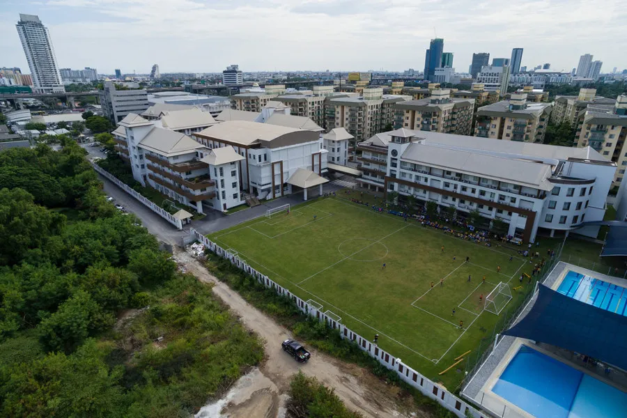 Aerial view of a school with sports fields, a soccer game in progress, and a swimming pool, set against a cityscape background.