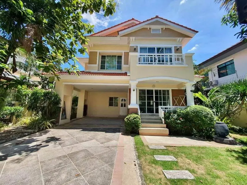 Two-story beige house with a red roof, surrounded by greenery, featuring a driveway and a small garden in front.