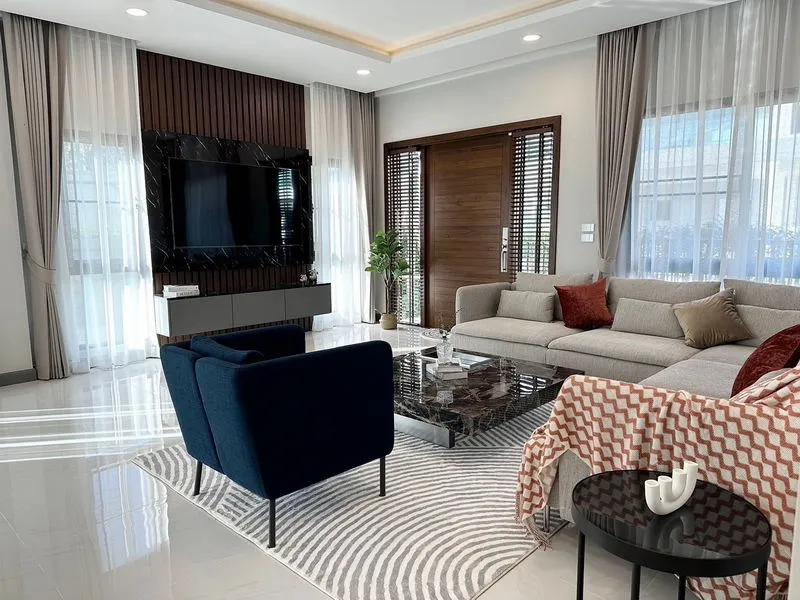 Modern living room with a gray sectional sofa, blue armchair, marble coffee table, and a wall-mounted TV. Natural light filters through sheer curtains, highlighting a stylish décor.