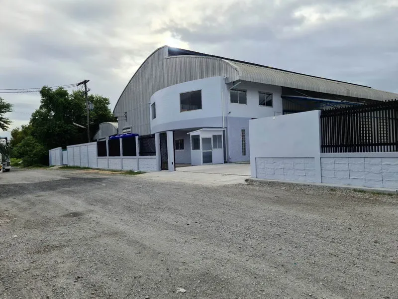 Industrial building with a curved roof, surrounded by a white fence and gate, situated on a gravel road under a cloudy sky.