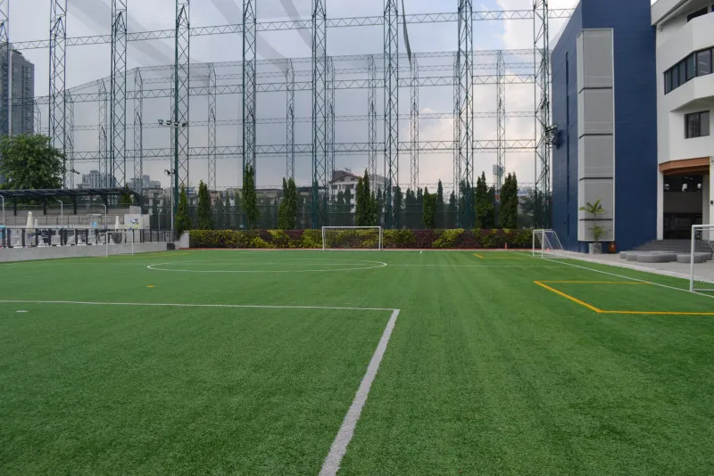 An empty artificial turf soccer field surrounded by buildings and high netting under a cloudy sky.