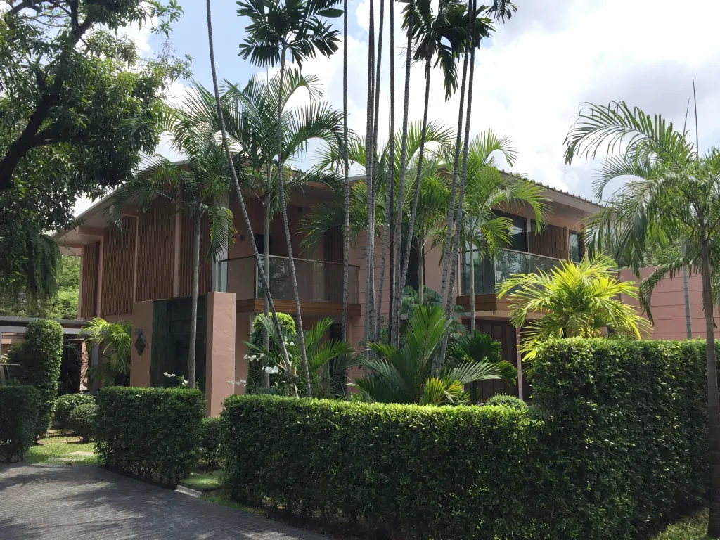 A modern two-story house surrounded by lush greenery and tall palm trees against a cloudy sky.