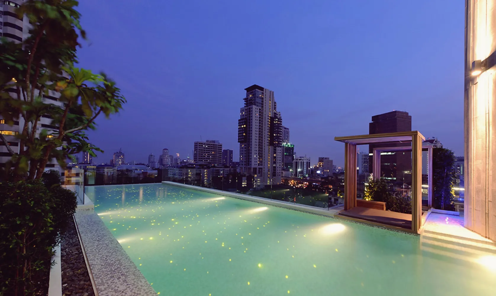Rooftop infinity pool with city skyline view at dusk, surrounded by buildings and lush greenery.