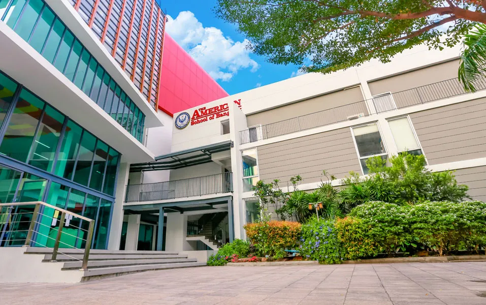 อาคารเรียนที่ทันสมัย ​​มีหน้าต่างกระจกบานใหญ่ ผนังสีเทา และสวนหย่อมที่จัดภูมิทัศน์อย่