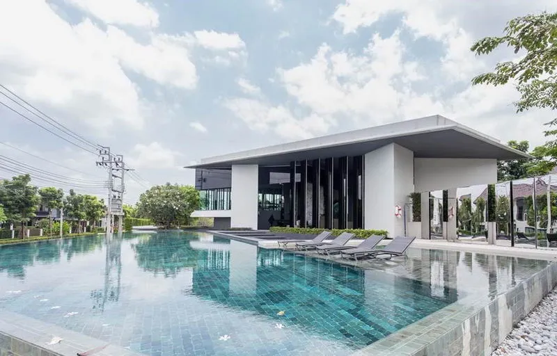 Modern building with large windows next to an outdoor pool, surrounded by greenery under a partly cloudy sky.