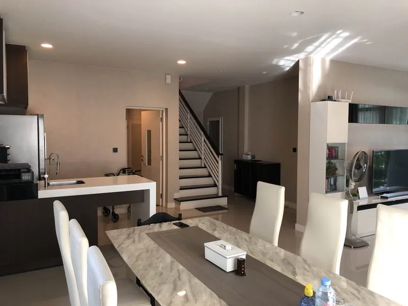 Modern dining and living area with marble table, white chairs, kitchen, stairs, and shelves.