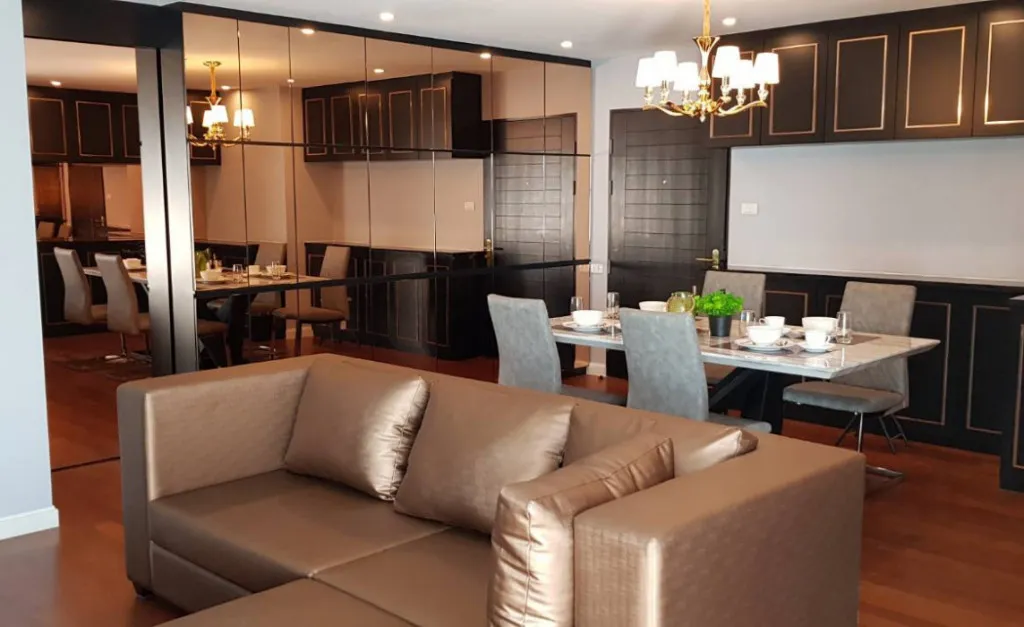 Elegant living and dining area with a brown sofa, marble dining table, gray chairs, and chandeliers, featuring dark wood cabinetry and mirrored walls.
