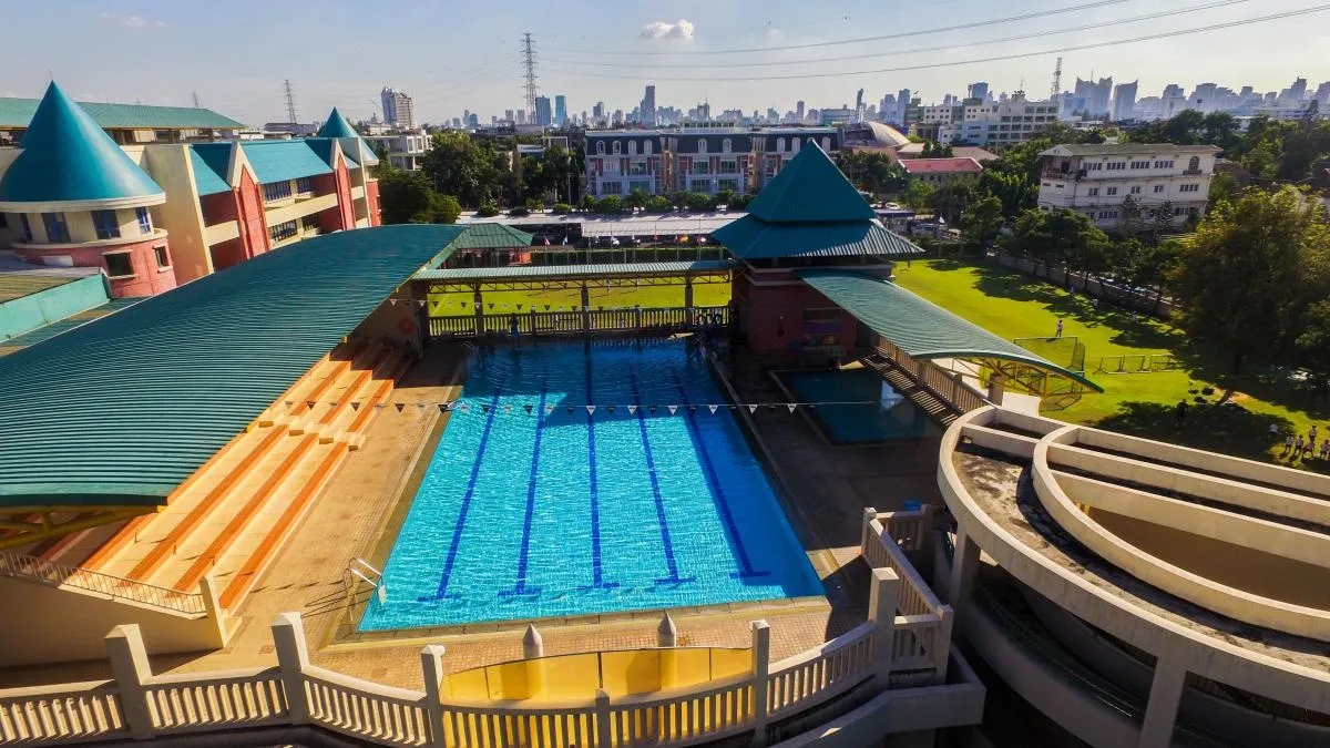 Aerial view of an outdoor swimming pool with lanes, surrounded by bleachers and buildings, and a city skyline in the background.