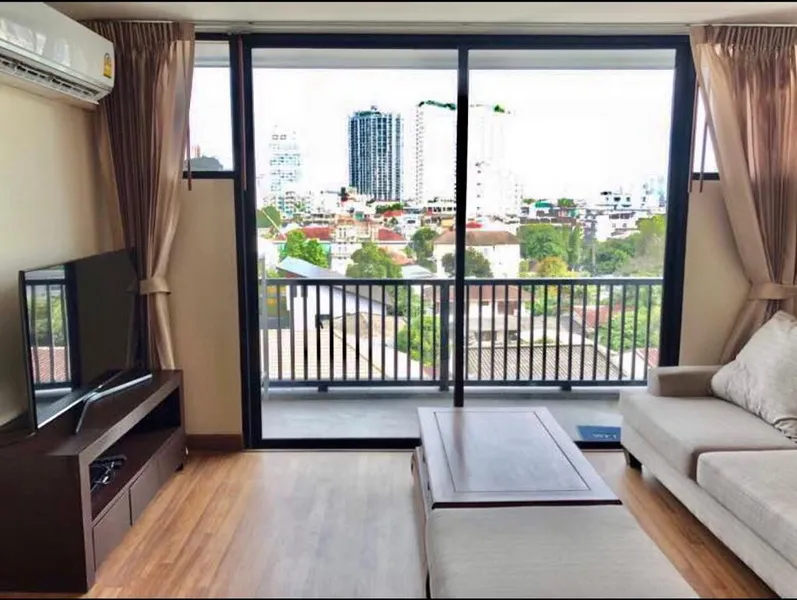 Modern living room with a beige sofa, flat-screen TV, wooden coffee table, and large sliding glass doors leading to a balcony with a cityscape view.