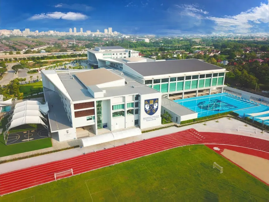 Aerial view of a modern school campus with large buildings, a swimming pool, sports courts, running tracks, and green fields, set against a cityscape background.