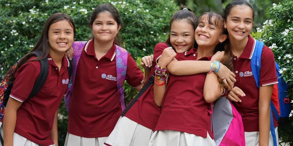 Group of five smiling schoolchildren in maroon uniforms with backpacks, standing outdoors with greenery in the background.