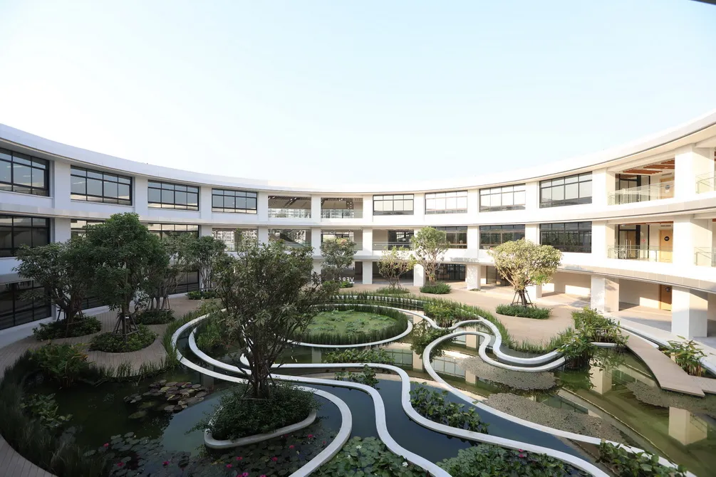 Modern building courtyard with curved walking paths and lush greenery surrounding a central pond under a clear blue sky.