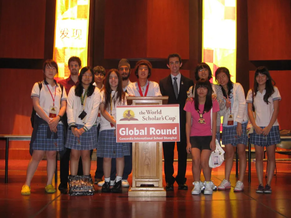 A group of students and adults stand on stage at a World Scholar's Cup Global Round event, wearing casual and school attire, smiling for a photo.
