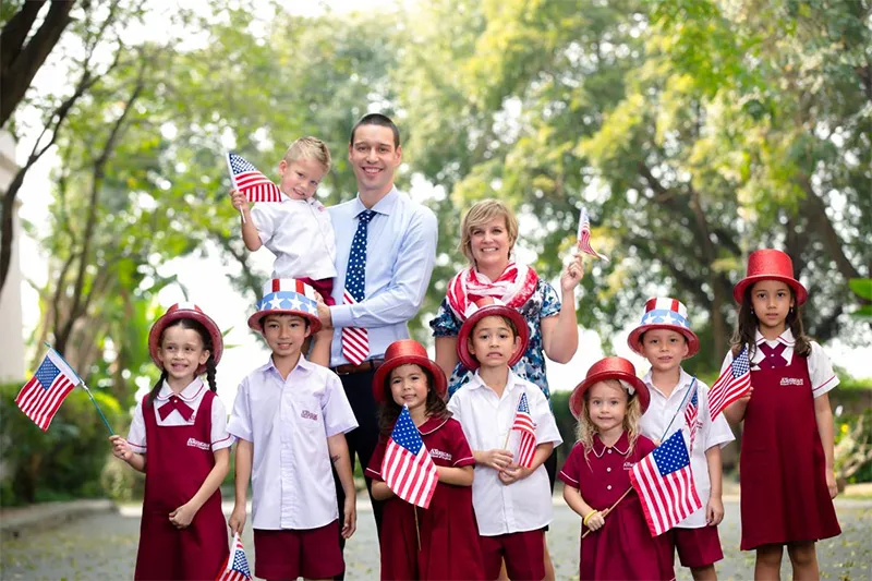 A group of children and two adults wearing red, white, and blue attire, holding American flags, standing outdoors with trees in the background.