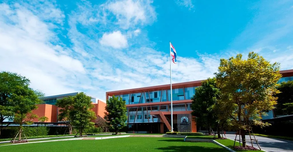 อาคารสมัยใหม่ที่มีหน้าต่างบานใหญ่ ล้อมรอบด้วยสนามหญ้าและต้นไม้เขียวขจี มีเสาธงชาติโบ