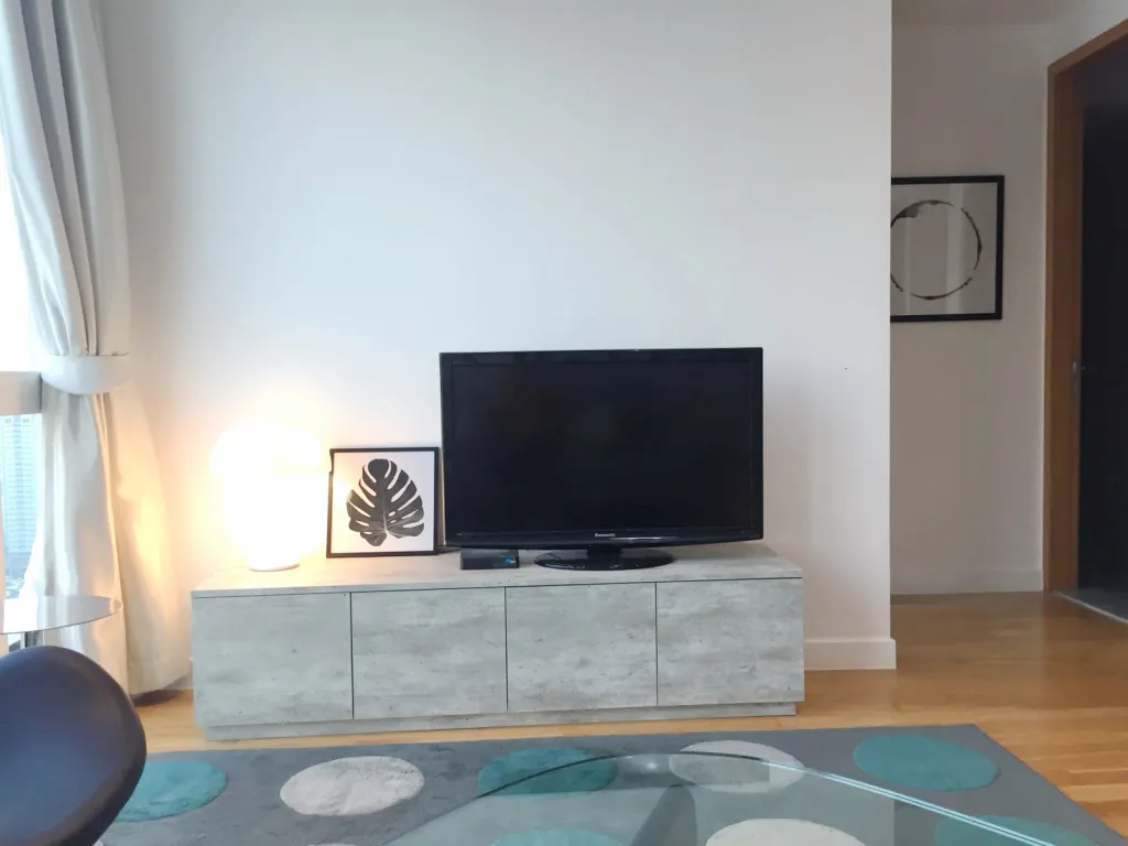 Modern living room with a TV on a light wood console, large floor lamp, framed leaf artwork, circular wall art, glass table, and patterned rug.