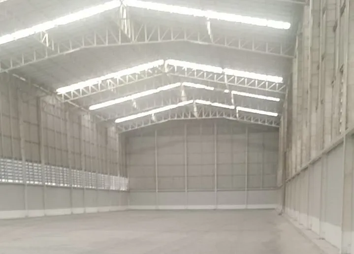 Large empty warehouse interior with high ceilings and steel framework, illuminated by natural light through translucent panels.