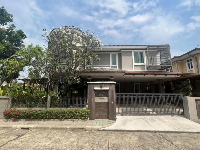Two-story house with a gated driveway, modern facade, and surrounding greenery under a partly cloudy sky.