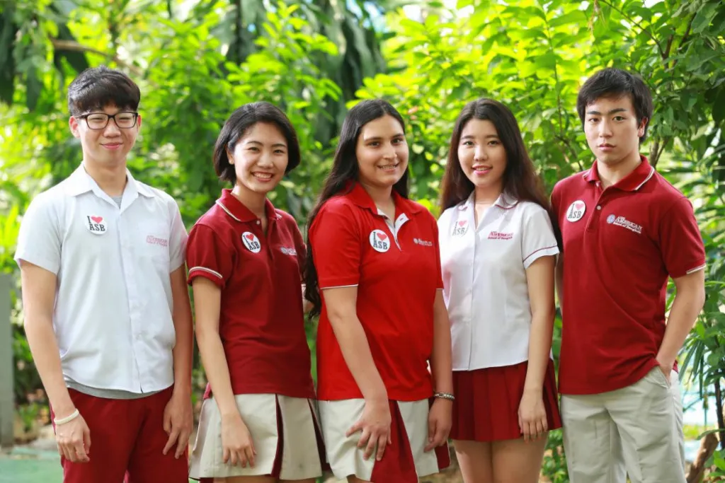Five students in uniform pose outdoors in front of greenery, wearing red and white shirts with the ASB logo.