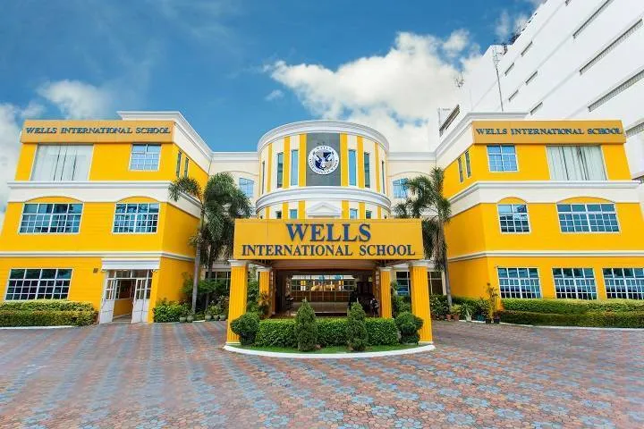 Yellow and white building of Wells International School with large entrance and sign, surrounded by palm trees under a blue sky.