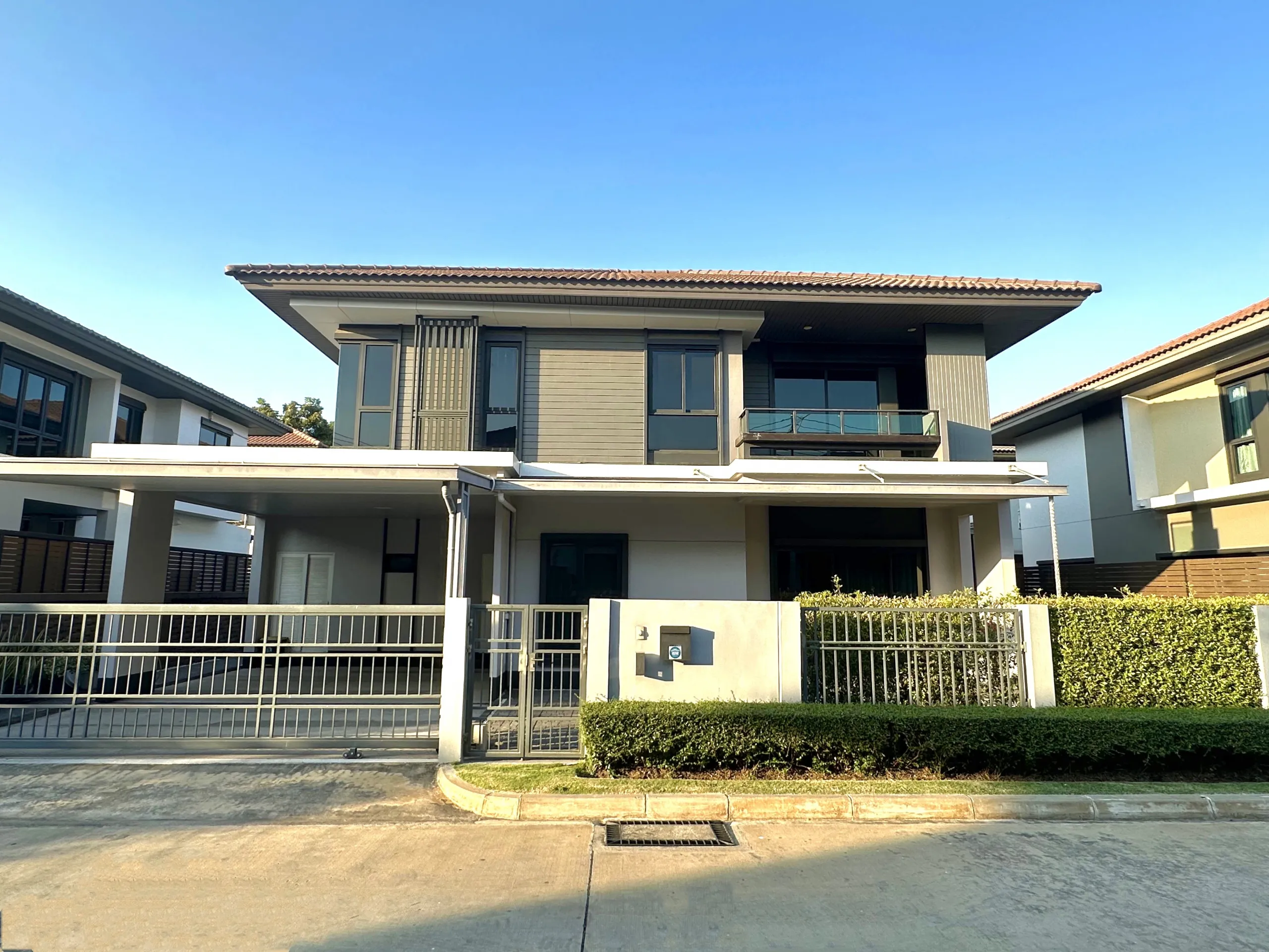 Modern two-story house with a flat roof, large windows, and a gated driveway, surrounded by hedges under a clear blue sky.