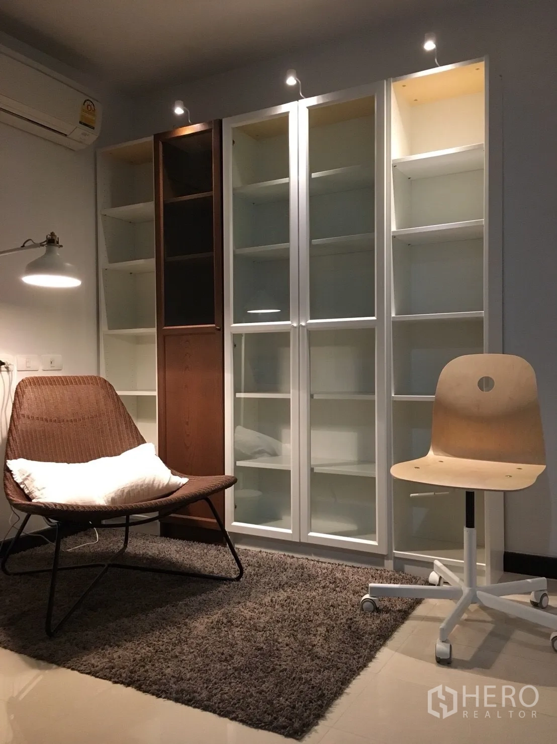 Condominium for rent in Watthana, Bangkok - Cozy reading nook with rattan lounge chair, rug, and tall glass-door cabinets under spotlights.