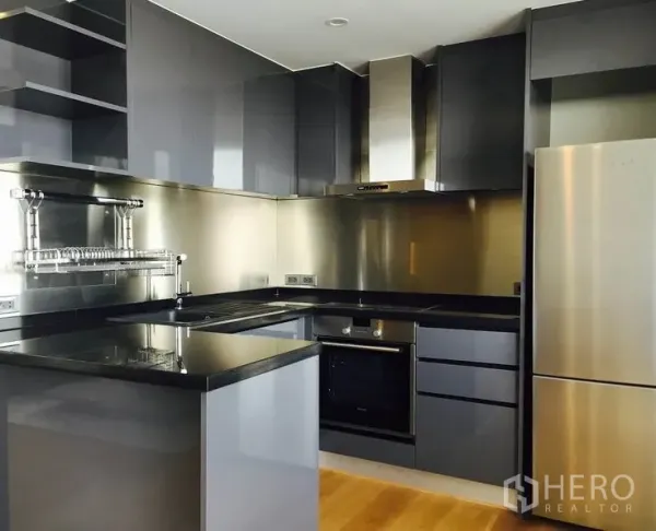 Condominium for rent in Watthana, Bangkok - Modern grey kitchen with L-shaped counters, stainless backsplash, built-in oven, range hood and full-size fridge.