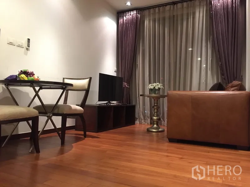 Condominium for rent in Khlong Toei, Bangkok - Warm living room with leather sofa, TV console, small dining set and floor-to-ceiling curtains on wooden flooring.