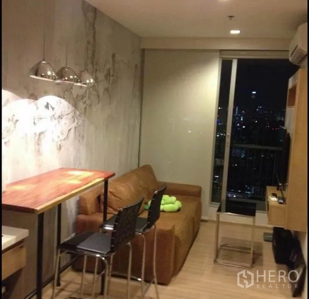 公寓 出售在Phaya Thai, Bangkok - 温馨客厅，棕色沙发与吧台及吊灯，面对落地窗欣赏夜景天际线。