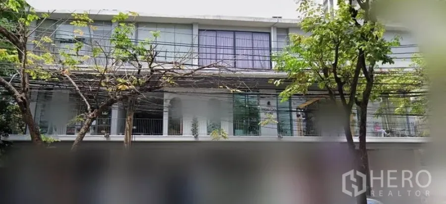 办公空间 出租在Khlong Toei, Bangkok - 低层建筑外观，大幅玻璃窗与阳台护栏，街道行树，位于素坤逸区域。