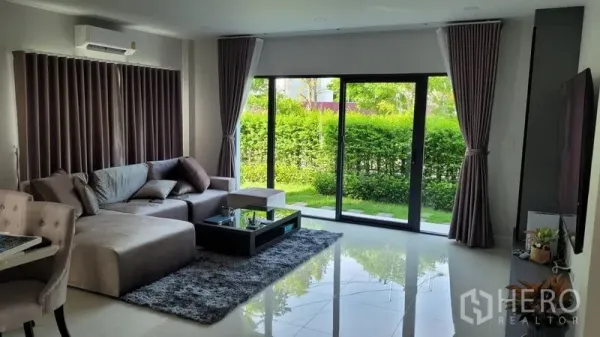 Detached House for rent in Don Mueang, Bangkok - Airy living room with L-shaped sofa and floor-to-ceiling sliding doors opening to a green garden.
